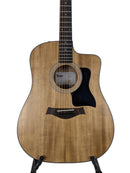 A Taylor acoustic-electric guitar with a natural wood finish and black pickguard, displayed on a stand against a light gray background.