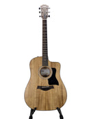 A Taylor acoustic-electric guitar with a natural wood body and black pickguard, displayed on a stand against a light gray background.