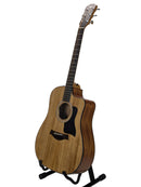 A Taylor acoustic-electric guitar with a natural wood body and dark pickguard, displayed on a black stand against a light gray background.
