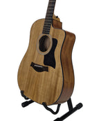 Taylor acoustic-electric guitar with a natural wood body and black cutaway body, displayed on a black stand against a white studio background.