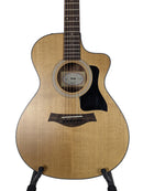 Taylor acoustic guitar with a natural wood finish and black pickguard, shown on a stand against a light gray background.