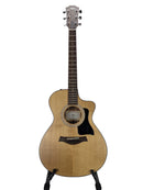 A Taylor acoustic-electric guitar with a natural wood finish and black pickguard, displayed on a stand against a light gray background.