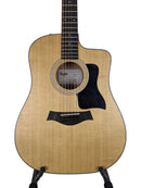 A Taylor acoustic-electric guitar with a natural wood finish and black pickguard, displayed on a stand against a light gray background.