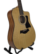 A Taylor acoustic-electric guitar with a natural wood top and dark brown sides, displayed on a black stand against a light gray background.