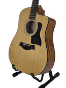 An acoustic-electric guitar with a natural wood top and dark brown sides, displayed on a black stand against a light gray background.