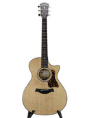 A Taylor acoustic-electric guitar with a natural wood body, black pickguard, and dark fretboard, displayed on a stand against a light gray background.