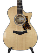 Taylor acoustic-electric guitar with a natural flamed maple top, black cutaway body contour, and black pickguard, displayed on a stand against a light gray background.