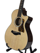 A Taylor acoustic-electric guitar with a natural wood top and dark brown body, displayed on a black stand against a light gray background.
