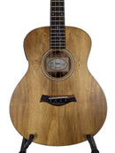 A wooden acoustic guitar with a natural light brown finish and a black neck, displayed on a stand against a light gray background.