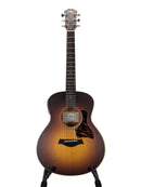 A Taylor acoustic guitar with a natural-to-brown sunburst finish, displayed on a stand against a light gray background.